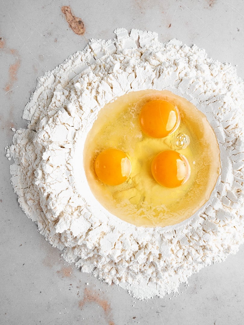 Three eggs cracked into the center of a flour well to make fresh homemade pasta dough.