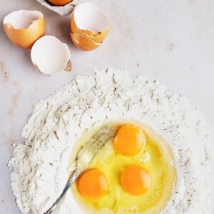 Three eggs cracked into the center of a flour well to make fresh homemade pasta dough with carton of eggs and a fork next to it.