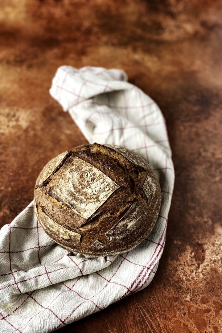 A whole wheat sourdough loaf with a white tea towel on a copper colored backdrop.