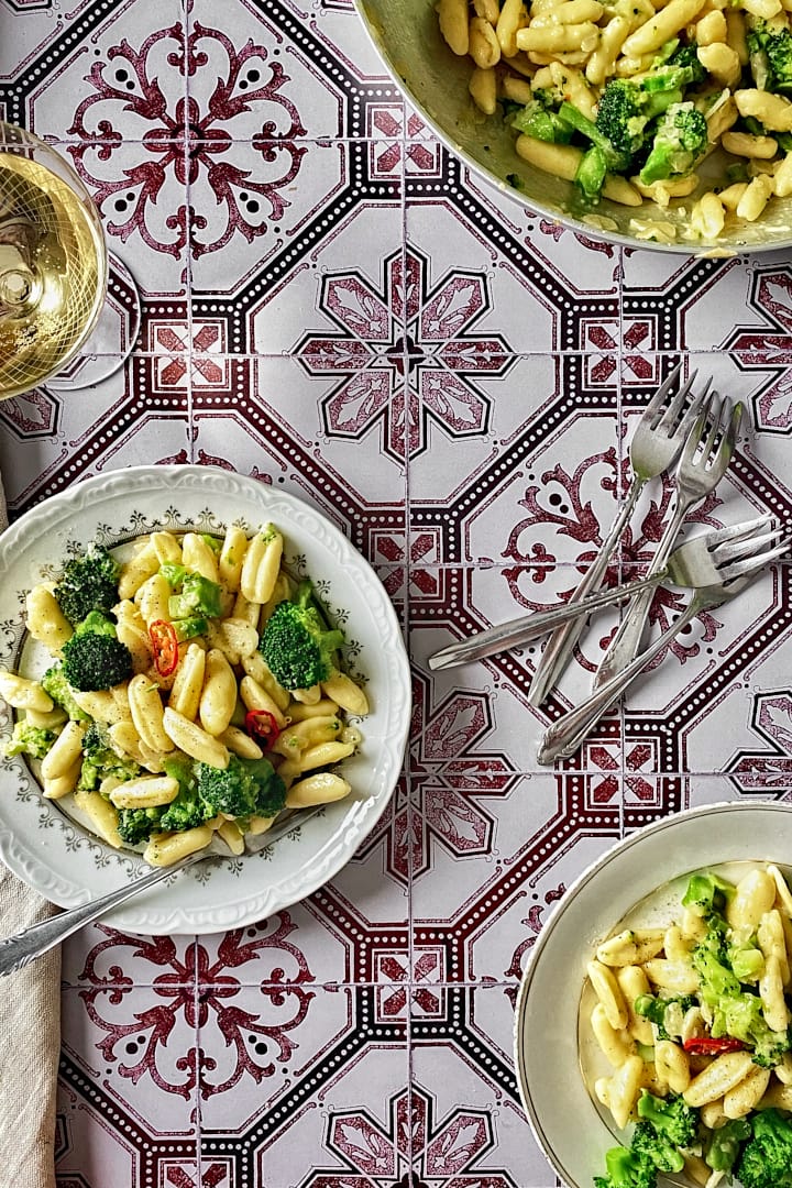 A pan and two plates of homemade cavatelli and broccoli pasta on a patterned tiles backdrop.