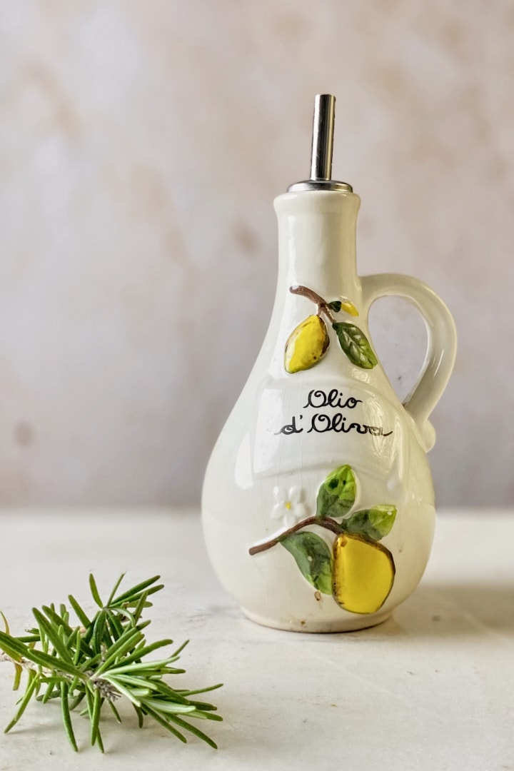A small bottle of olive oil and stalk of fresh rosemary in front of a neutral marble backdrop.