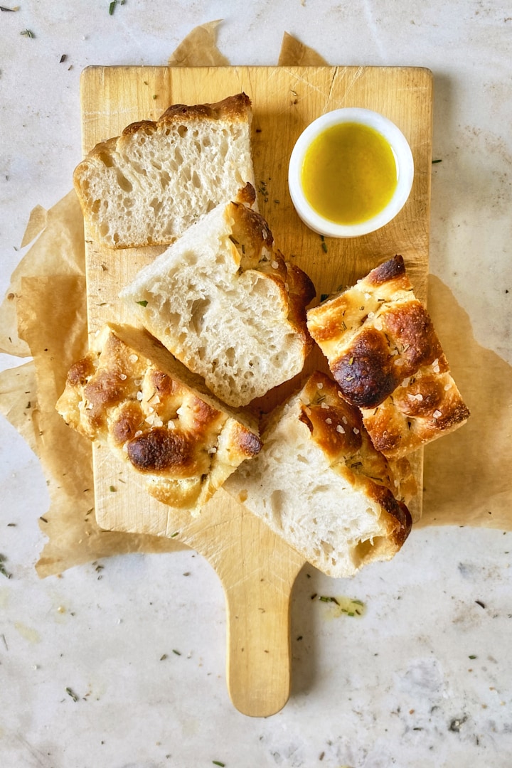 Pieces of homemade focaccia on a wooden board with a small bowl of olive oil for dipping.