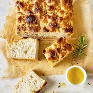 Flatlay of a freshly baked focaccia on a piece of parchment paper with fresh rosemary and olive oil on the side.