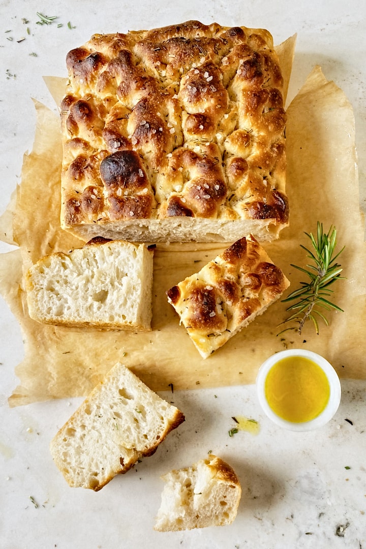 Flatlay of a freshly baked focaccia on a piece of parchment paper with fresh rosemary and olive oil on the side.