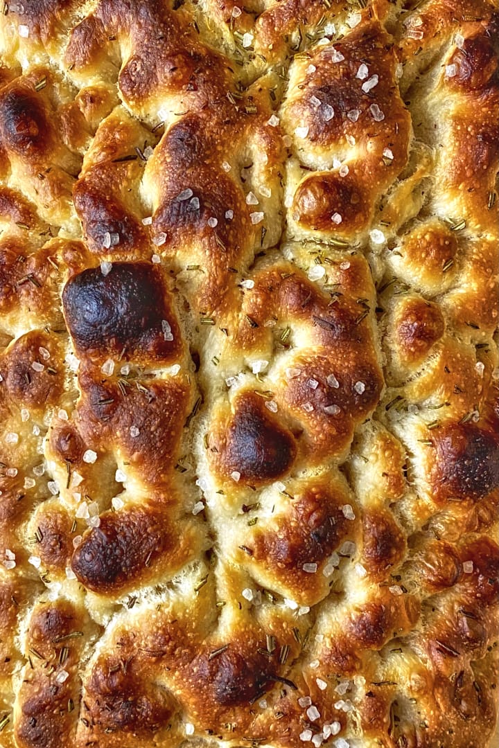 Close-up of the bubbly surface of a homemade focaccia with rosemary and flakey sea salt.