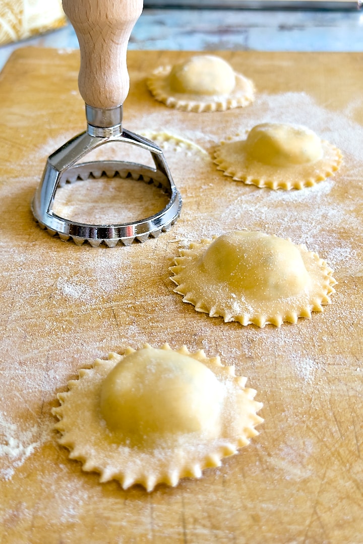Fresh ravioli on a wooden board with a ravioli stamp next to them.