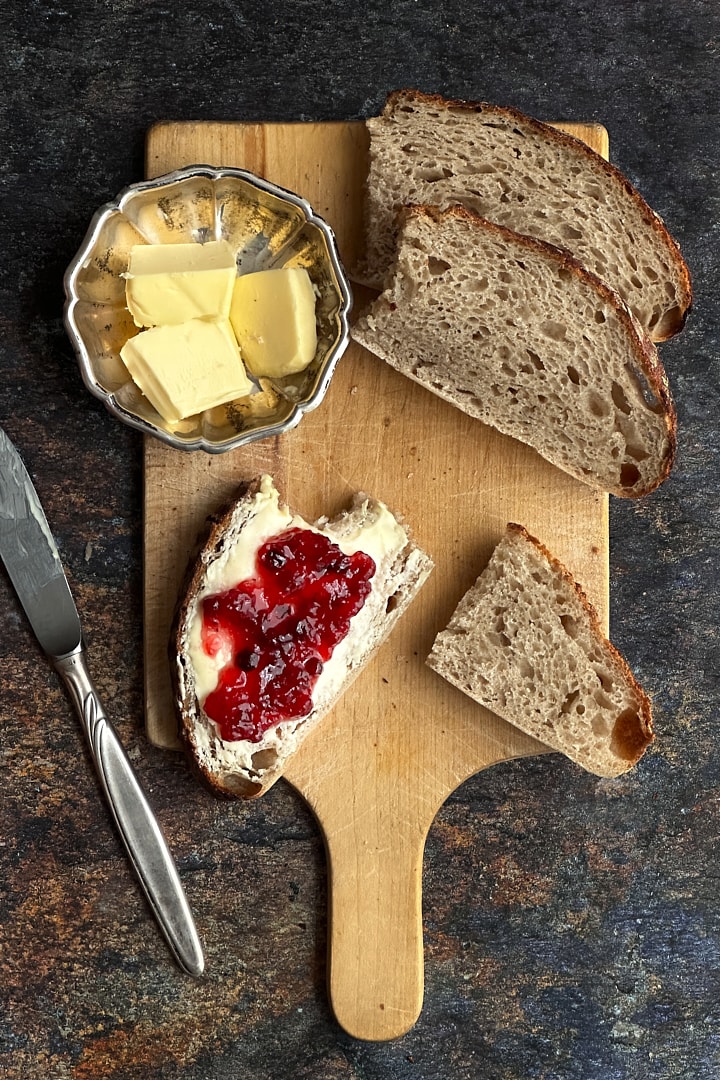 Slices of rustic sourdough bread on a wooden board with butter and jam.