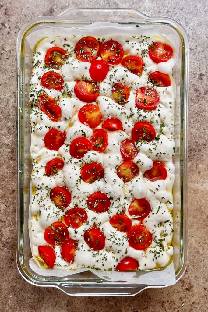 A baking dish with bubbly focaccia dough topped with cherry tomatoes and chopped rosemary. .