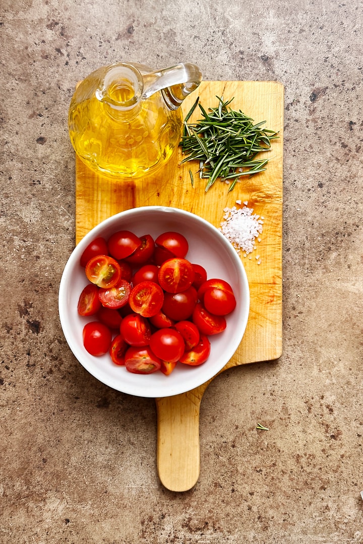 A wooden board with the toppings for homemade focaccia: olive oil, flakey sea salt, fresh rosemary and cherry tomatoes.