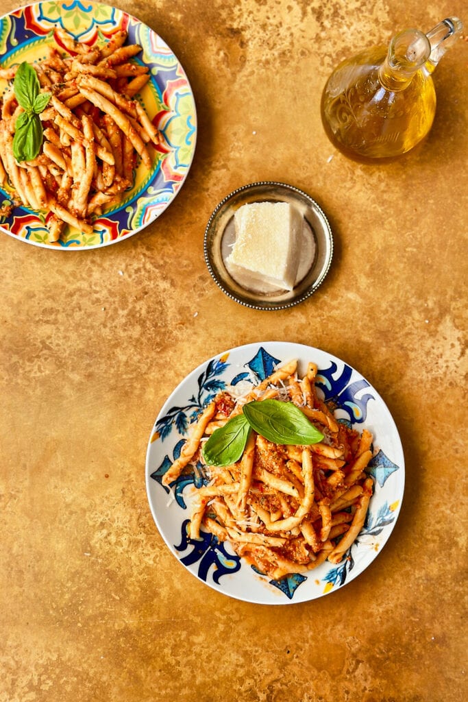 a flatlay table scene featuring two plates of pasta with pesto alla trapanese, pecorino cheese, and a small jug of olive oil on a terracotta backdrop.