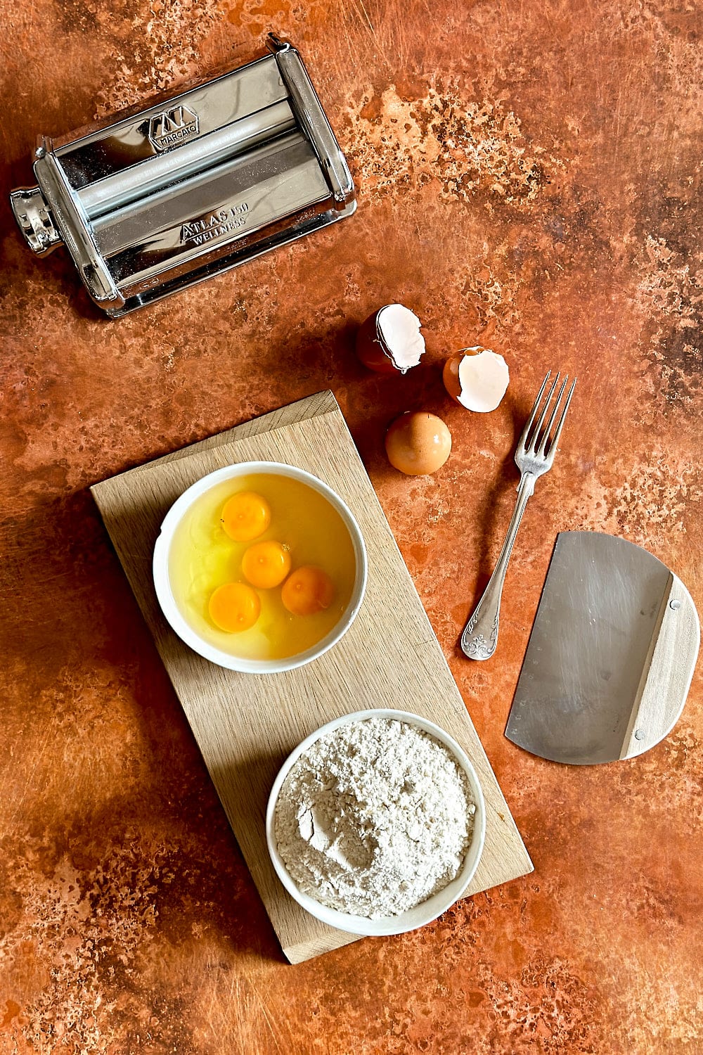 The tools and ingredients needed to make homemade tagliatelle: Flour, eggs, bench scraper, fork and pasta machine.