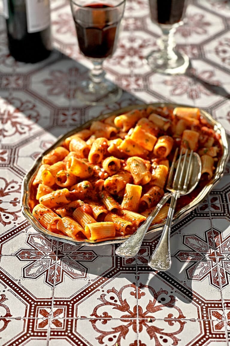 Two glasses of red wine and a plate of rigatoni arrabbiata on a patterned tile backdrop.