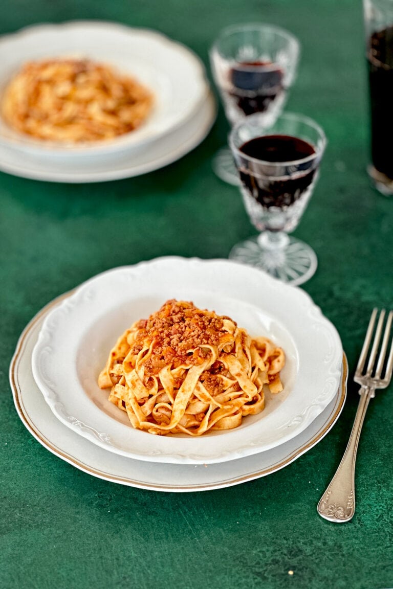 Two white plates with homemade tagliatelle alla bolognese on a dark green table.