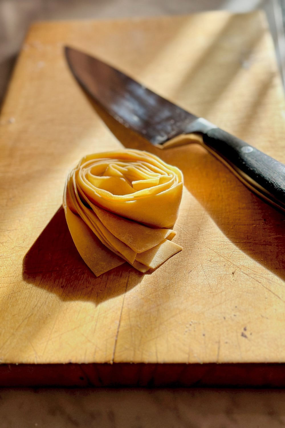 A pappardelle pasta nest next to a sharp knife on a wooden board.