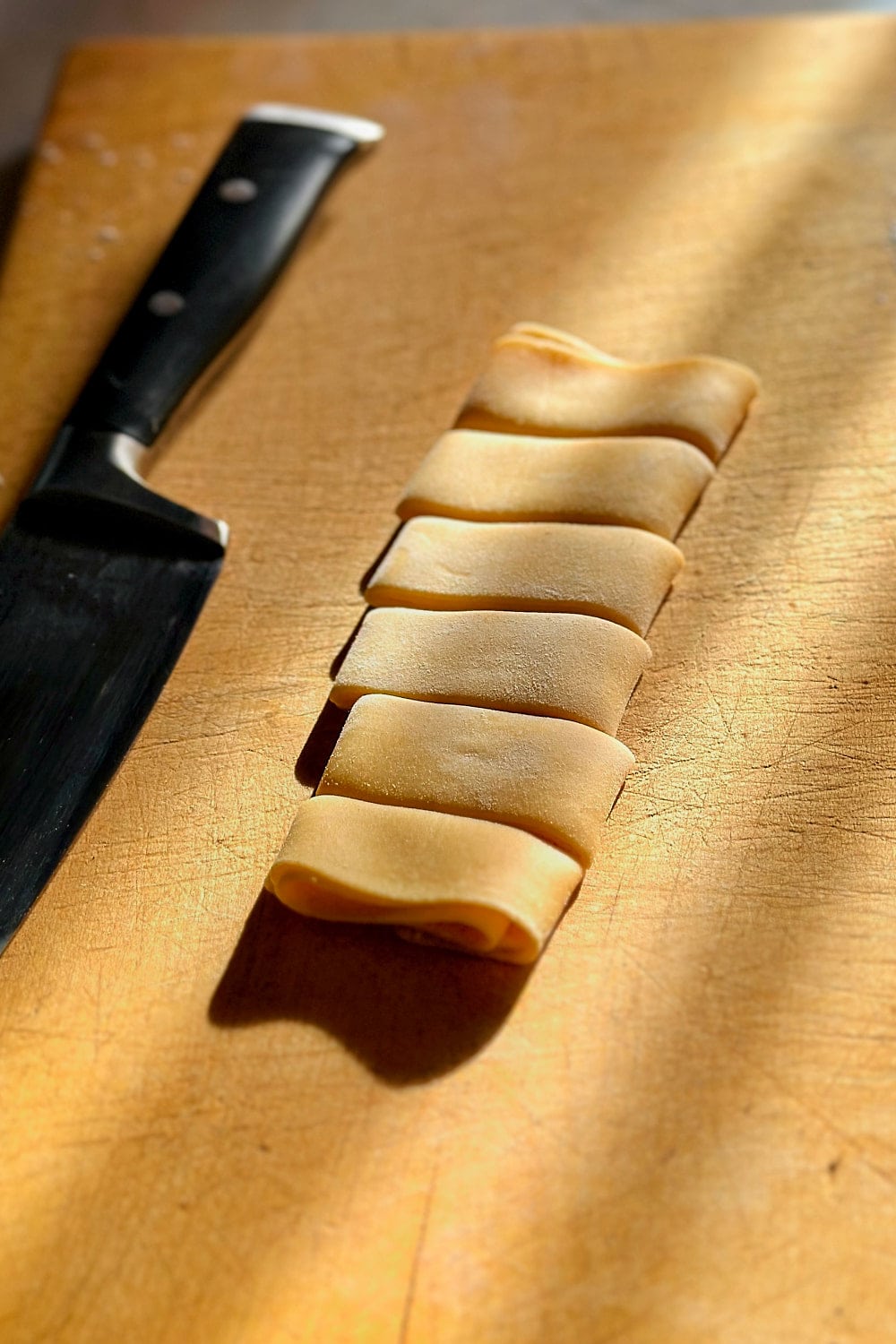Cutting a sheet of pasta dough into wide strips on a wooden board to make homemade pappardelle pasta.