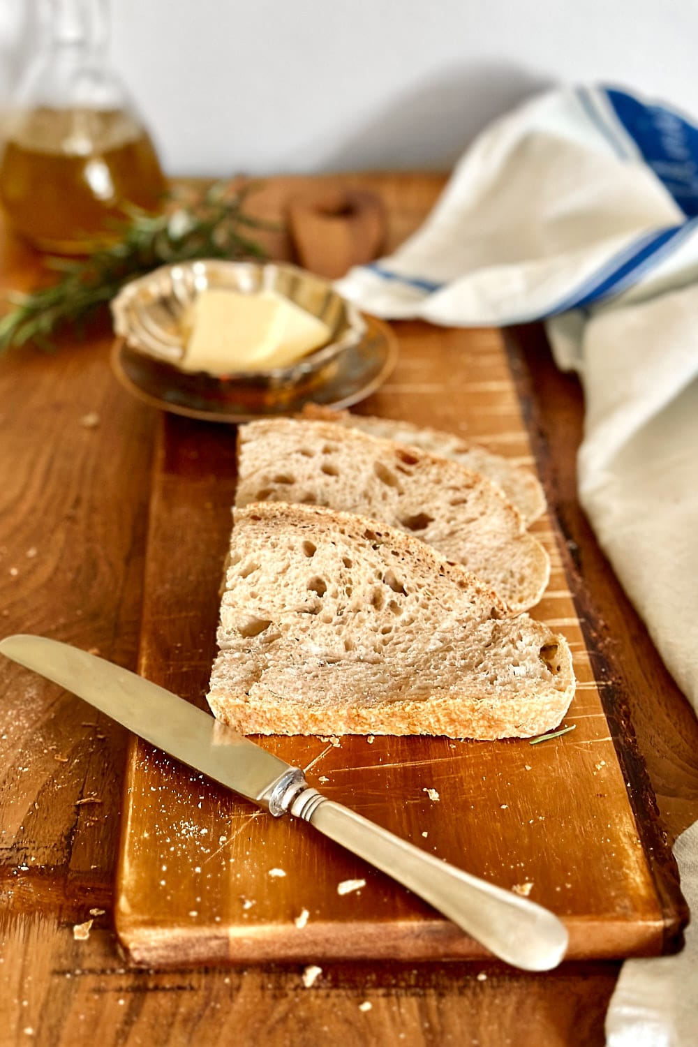Three slices of rosemary sourdough bread on a wooden board with some butter and a knife next to it.