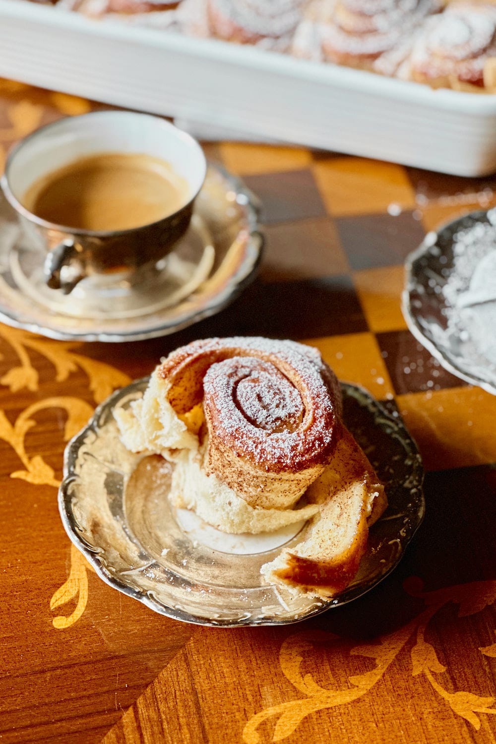 A sourdough cinnamon bun dusted with powdered sugar and a cup of coffee next to it.