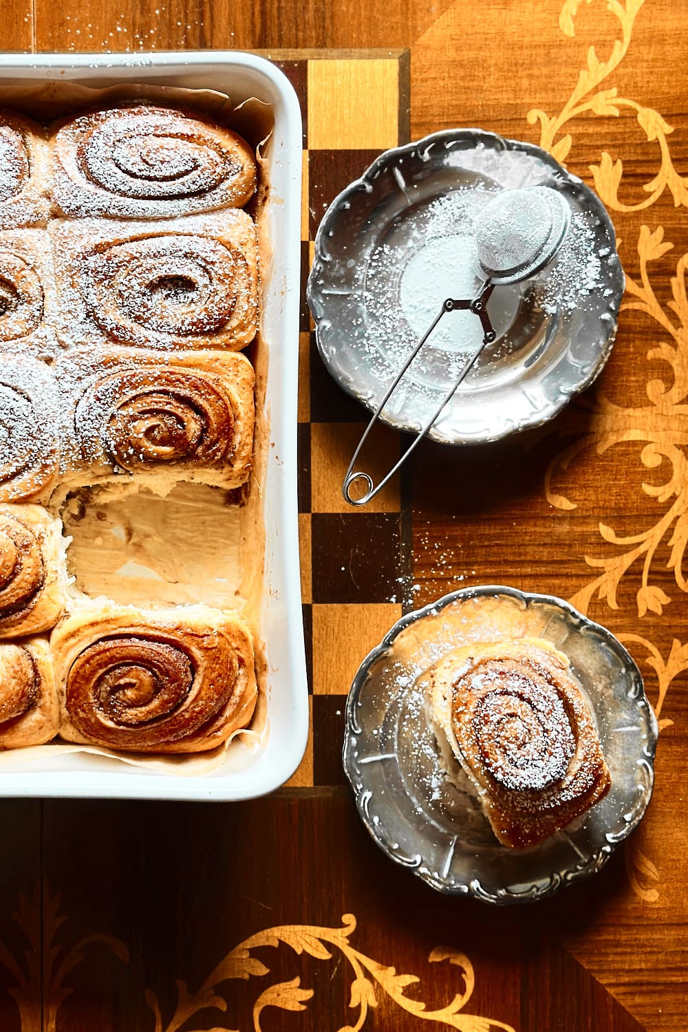 A baking dish with sourdough cinnamon buns and one bun on a small separate plate, dusted with powdered sugar.