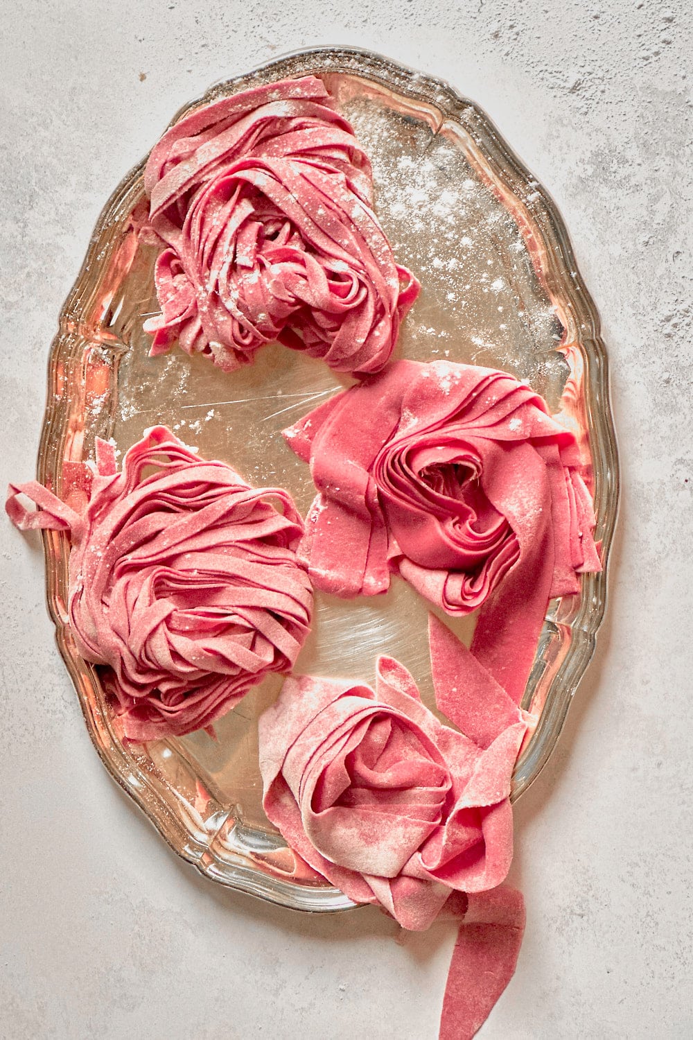 Homemade pink beet pasta nests on a silver tray on a light grey backdrop.